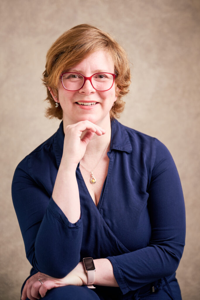 A professional picture of a woman with short red hair and a blue shirt dress sitting with her hand tucked under her chin